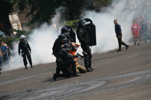 Eine Gruppe von Menschen steht auf einer Straße, einige tragen Helme und halten Waffen, andere halten verschiedene Gegenstände, während im Hintergrund Rauch aufsteigt, umgeben von Pflanzen, Bäumen, Gebäuden und anderen Gegenständen, während ein Mann von Polizisten in Einsatzausrüstung eskortiert wird.