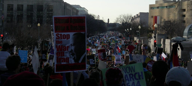 Menschen protestieren auf einer Straße und halten Plakate in den Händen, im Hintergrund sind Gebäude, Bäume, Laternenpfähle und der Himmel zu sehen.
