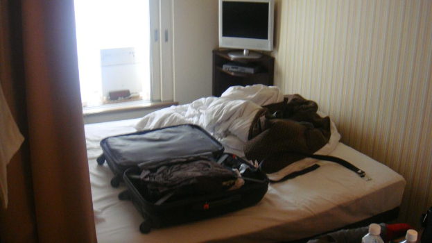 Ein Bett mit Gepäckkoffern und Kleidern darauf, mit einem Fernseher auf einem Tisch im Hintergrund und einer Wand.