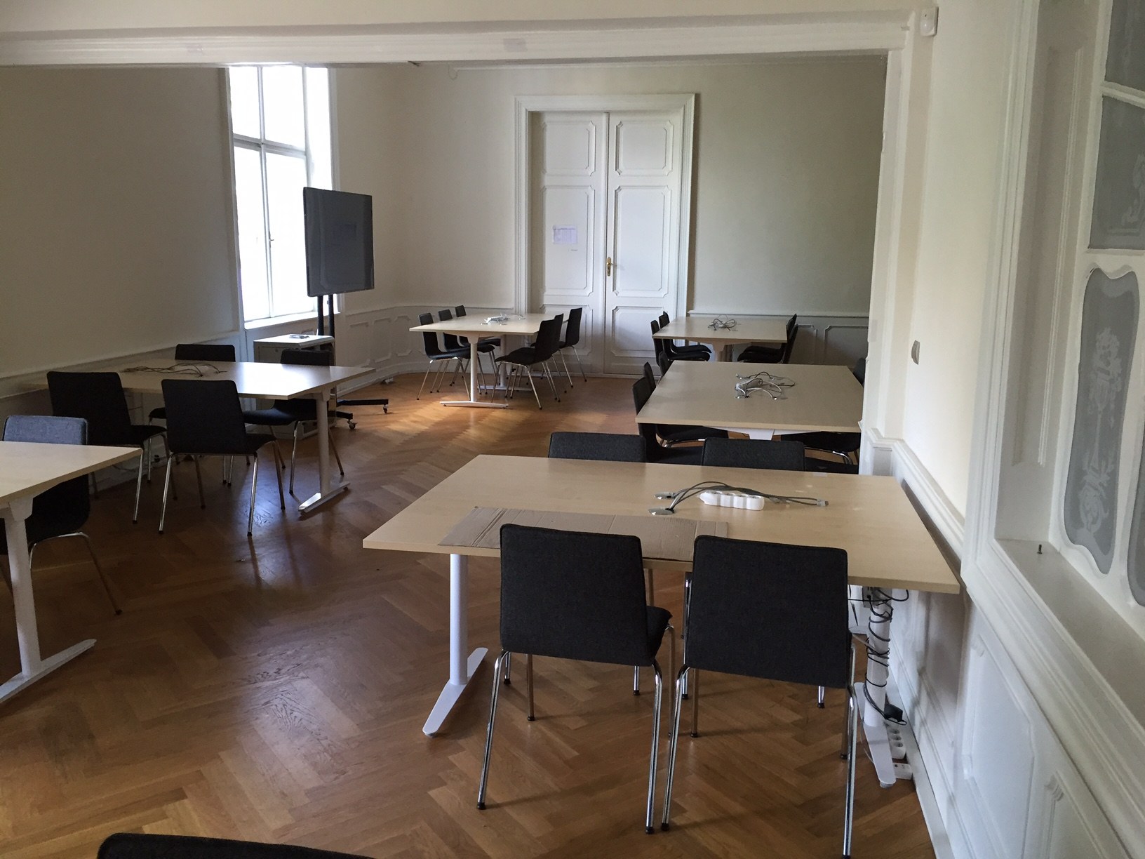 Großer Konferenzraum mit Tischen, Stühlen, einem Fernseher auf einem Ständer, einer Tür, einem Fenster und einer Wand, verfügbar zur Miete in Paris.
