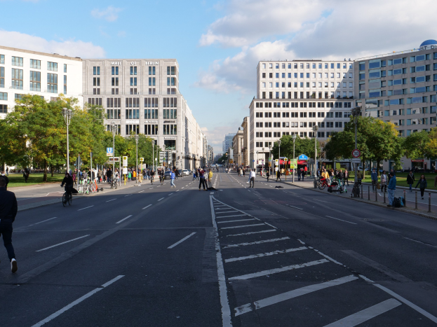 Eine belebte Straßenkreuzung in Berlin, Deutschland, mit Fußgängern und Radfahrern auf der Straße, umgeben von hohen Gebäuden, Bäumen, Laternen und Schildern unter einem bewölkten Himmel.