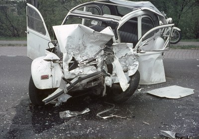 Ein schwer beschädigt aussehendes Auto mit zertrümmerter Front steht am Straßenrand, mit Bäumen und einem Gebäude im Hintergrund und einem weiteren Fahrzeug in der Ferne.