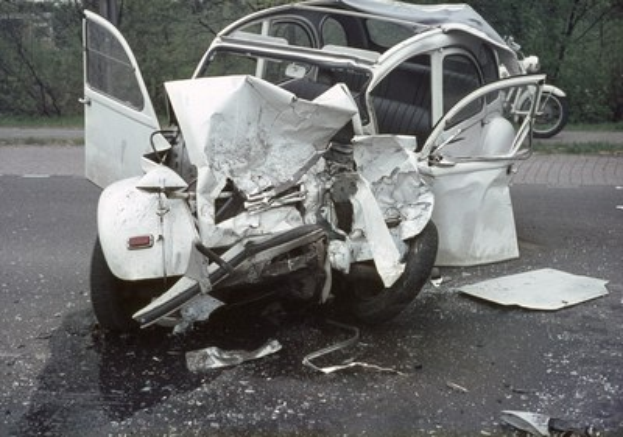 Ein schwer beschädigt aussehendes Auto mit zertrümmerter Front steht am Straßenrand, mit Bäumen und einem Gebäude im Hintergrund und einem weiteren Fahrzeug in der Ferne.