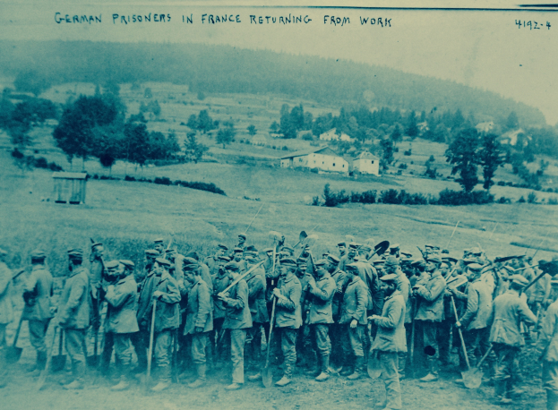 Eine Gruppe deutscher Gefangener in Frankreich mit Waffen auf dem Boden stehend, umgeben von Bäumen, Häusern und einem klaren blauen Himmel.
