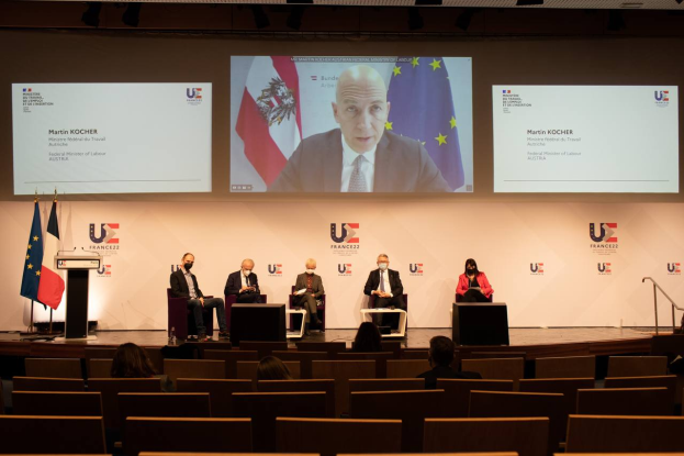 Eine Gruppe von Menschen sitzt auf einer Bühne in einem Halbkreis um ein Podium mit Mikrofon, mit Flaggen auf beiden Seiten, einem Bildschirm mit Text und Bildern im Hintergrund und Scheinwerfern, die die Szene beleuchten, wahrscheinlich bei einer Pressekonferenz der Europäischen Union.