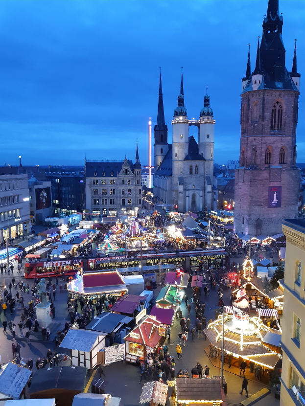 Ein nächtlicher Weihnachtsmarkt in Köln, Deutschland, mit Menschen auf dem Boden, die von den Lichtern der Stände und Gebäude beleuchtet werden, unter einem bewölkten Himmel.