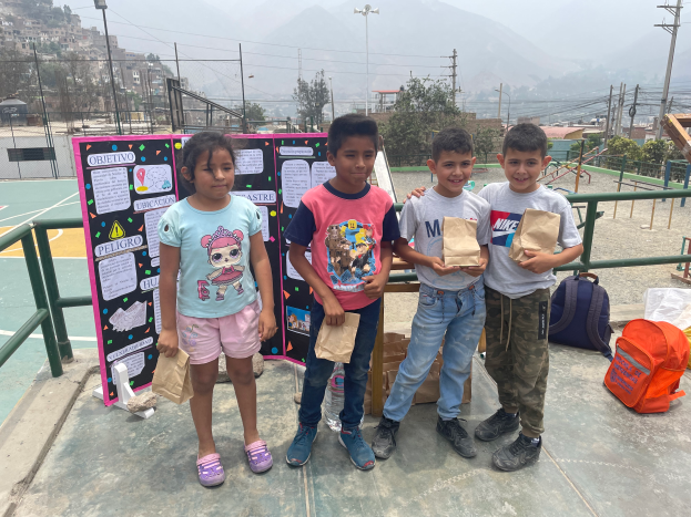 Eine Gruppe von Kindern steht auf einem Basketballfeld, jedes hält eine Papiertüte, mit Tüten und Gegenständen rechts und einer Tafel mit Text links, mit Zäunen, Pfosten, Straßenlaternen, Bäumen, Gebäuden, Hügeln und Himmel im Hintergrund.