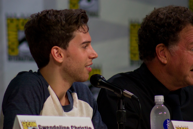 Ein Mann, der vor einem Mikrofon lächelt, mit zwei Namensschildern und einer Flasche sowie einem Banner im Hintergrund.