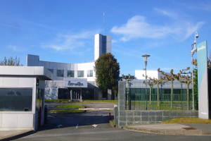 Ein großes, mehrfenstriges Gebäude mit einem Tor davor, umgeben von Straßenmasten, Lampen, Gittern, Bäumen, Gras und einem bewölkten Himmel, das als Hauptquartier von Iberostica in den Niederlanden dient.