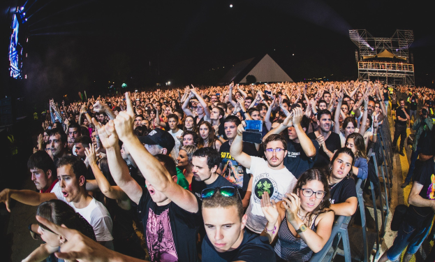 Eine große Menschenmenge auf einem Musikfestival mit erhobenen Händen, einige sitzen auf Stühlen und andere stehen, begrenzt von einem Metallzaun auf der rechten Seite, mit Gebäuden und Lichtern im Hintergrund unter einem dunklen Himmel.