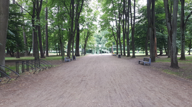 Ein Park mit einem Schotterweg, der von Bäumen und Bänken gesäumt ist und zu einem Gebäude unter einem klaren blauen Himmel führt.