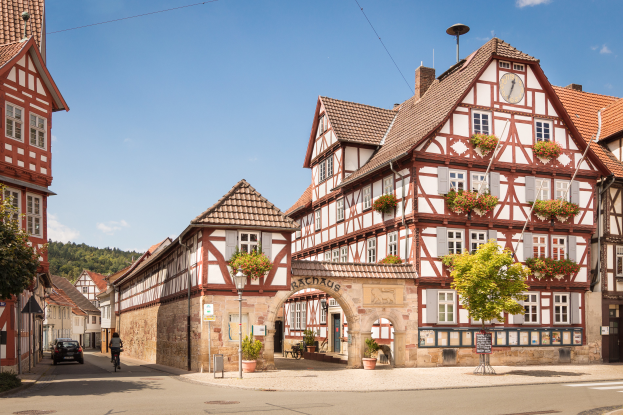 Eine Straße in Rothenburg ob der Tauber, Deutschland, mit Gebäuden, Fenstern, Bäumen, Laternenpfählen, einer Person auf einem Fahrrad und einer hügeligen Landschaft mit Wolken am Himmel im Hintergrund.