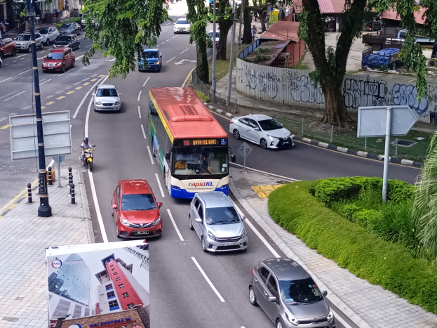 Eine belebte Stadtstraße mit zahlreichen Autos, Bussen und Motorrädern, umgeben von Pfählen, Schildern, Laternen, Bäumen, Pflanzen, Gras, Gebäuden und einer Wand mit Schrift.
