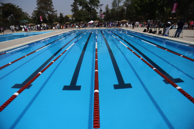 Großes Schwimmbecken mit Bahnentrennung, umgeben von Menschen mit Taschen und Gegenständen, Zelten, Bannern, Masten, einem Schild und einem bewölkten Himmel im Hintergrund.