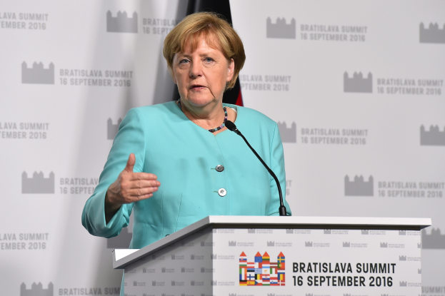 Bundeskanzlerin Angela Merkel hält eine Rede auf dem Bratislava-Gipfel, trägt ein blaues Kleid und steht an einem Podium mit zwei Mikrofonen, mit einem Banner und einer Flagge im Hintergrund.