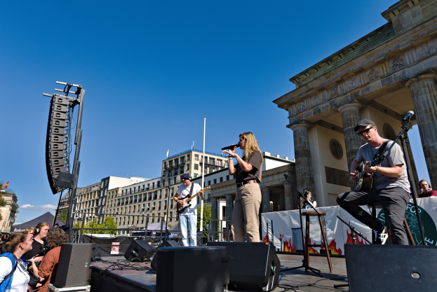 Eine Gruppe von Menschen, die auf einer Bühne vor dem Brandenburger Tor in Berlin Musik machen, mit Lautsprechern und Equipment im Bild, unter einem klaren blauen Himmel.