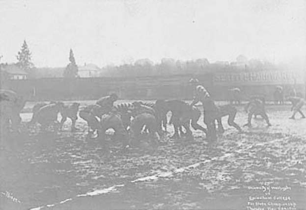 Ein Schwarz-Weiß-Foto einer Gruppe von Menschen, die auf einem Feld Fußball spielen, mit Pferden im Vordergrund und Bäumen, Gebäuden und Himmel im Hintergrund. Unten auf dem Bild steht der Text "1918-1918 Fußball an der Staatsuniversität".