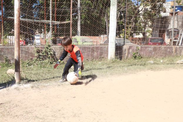 Ein junger Junge spielt Fußball auf einem Schmutzfeld mit einem bewölkten Himmel, umgeben von Gras, Pflanzen, Pfählen, einem Zaun, einer Wand, Bäumen, Fahrzeugen und Gebäuden, trägt Schuhe und ist aktiv im Spiel.
