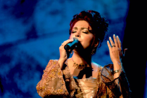 Eine Frau in einem goldenen Kleid singt in ein Mikrofon auf der Bühne der Eurovision Song Contest, mit einem blauen und schwarzen Hintergrund.