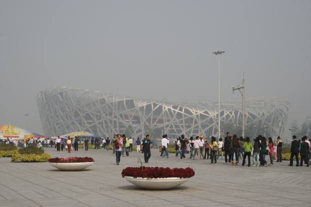 Olympiastadion in Peking, China mit Menschen, die herumlaufen, Blumenpflanzen im Vordergrund, Laternenmasten, Bäume und einen klaren blauen Himmel im Hintergrund.