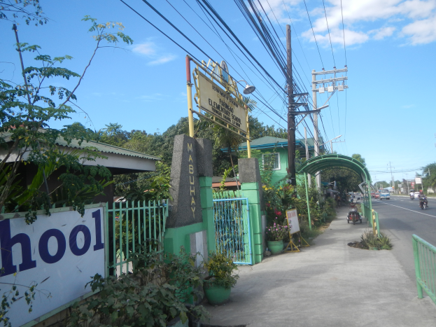 Der Eingang einer Schule mit einem Schild, auf dem "Schule" steht, an der Seite einer Straße, mit einem Bogen, einem Tor, einer Namenstafel, Pflanzen, Bäumen, Strommasten mit Drähten und einem bewölkten Himmel im Hintergrund.