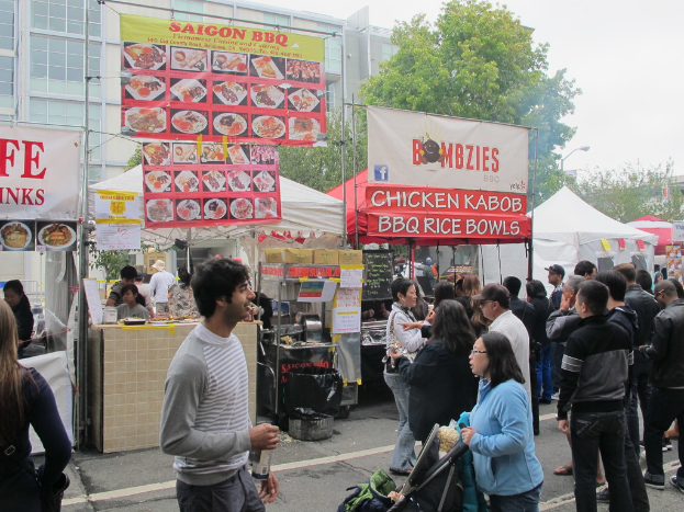 Menschen stehen auf einer Straße vor der Stadt, ein Mann in einem grauen und weißen T-Shirt hält eine Wasserflasche und eine Frau in einer blauen Jacke schiebt einen Kinderwagen. Im Hintergrund sind Stände mit weißen und roten Zelten, farbige Banner und Gebäude, Bäume und eine Straßenlaterne zu sehen.