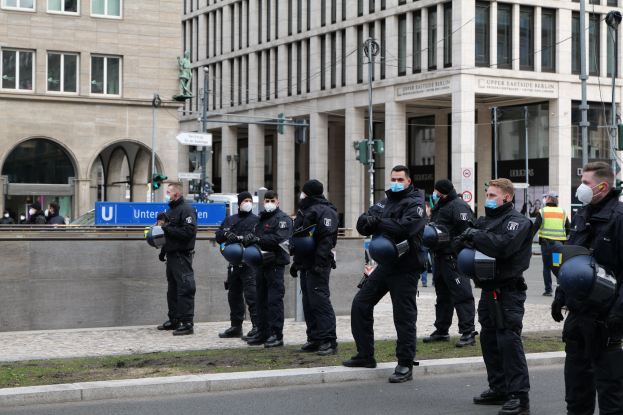 Eine Gruppe von Polizisten in schwarzen Uniformen und Masken steht vor einem Glasfenstergebäude mit Säulen in Berlin, Deutschland, einige halten Helme, mit Laternen, Verkehrszeichen, Texttafeln, einer Statue, anderen Menschen und grasbewachsenem Boden im Hintergrund.