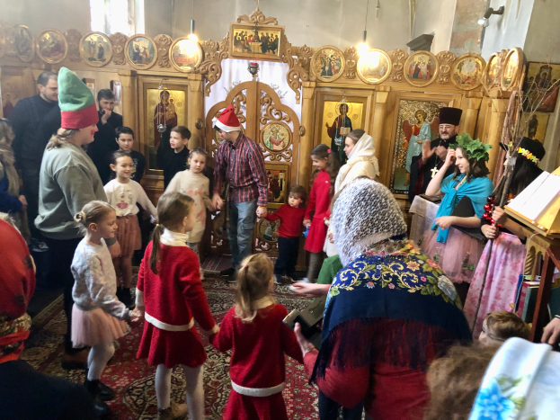 Eine Gruppe von Menschen steht vor einer Kirche, einige tragen Mützen, mit einem Buchständer rechts, Fotorahmen, Lichtern und einem Fenster im Hintergrund, während einer Weihnachtsfeier.