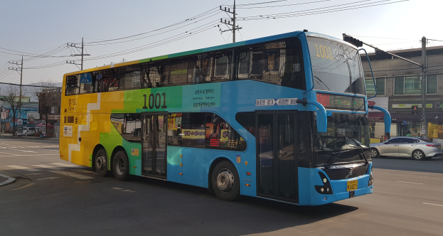 Ein blauer und gelber Doppeldeckerbus fährt auf einer Straße mit anderen Fahrzeugen, Strommasten, Gebäuden, Bäumen und einem klaren blauen Himmel im Hintergrund.