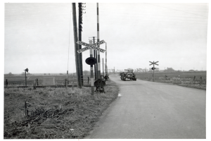 Ein Schwarz-Weiß-Foto eines Zuges, der eine Straße überquert, mit einem Fahrzeug, das daneben fährt, Pfählen mit Schildern auf beiden Seiten und Bäumen und einem klaren Himmel im Hintergrund.
