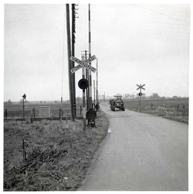 Ein Schwarz-Weiß-Foto eines Zuges, der eine Straße überquert, mit einem Fahrzeug, das daneben fährt, Pfählen mit Schildern auf beiden Seiten und Bäumen und einem klaren Himmel im Hintergrund.
