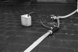 Schwarz-weiß-Foto eines Gasbehälters mit einem Schlauch, der vor einem Gebäude mit einer Tür im Hintergrund liegt.