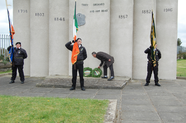 Drei Personen in Uniform stehen mit Flaggen verschiedener Farben, während ein Mann hinter ihnen grüne Kränze hält, vor einer Wand oder Säulen mit Text, Gras, Gehweg, einem Zaun auf der linken Seite und Bäumen und einem Gebäude auf der rechten Seite.