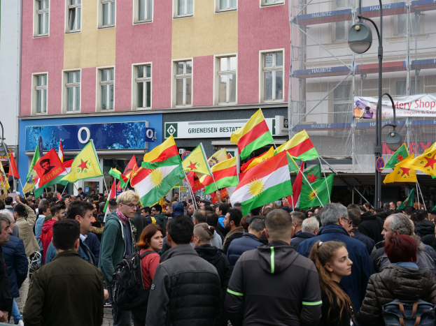 Große Gruppe von Menschen, die die Straße entlanggehen und kurdische Flaggen schwenken, mit Laternenmasten und Gebäuden an der Straße und Texttafeln im Hintergrund.
