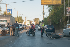 Eine Gruppe von Menschen steht um ein verunglücktes Motorrad auf der Seite einer Straße mit mehreren Fahrzeugen, darunter ein Lastwagen, und Hintergrundelementen wie Bäumen, Pfählen, Lampen, Schildern und dem Himmel.