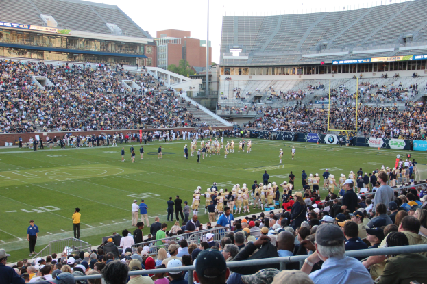 Ein Fußballspiel im Gange mit Spielern auf dem Feld und Zuschauern auf den Tribünen unter einem klaren blauen Himmel.