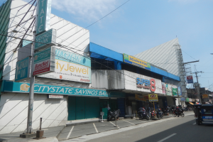 Eine belebte Stadtstraße in Cebu City mit Fahrzeugen, Fußgängern, Gebäuden und der Citystate Savings Bank als Mittelpunkt unter einem bewölkten Himmel.