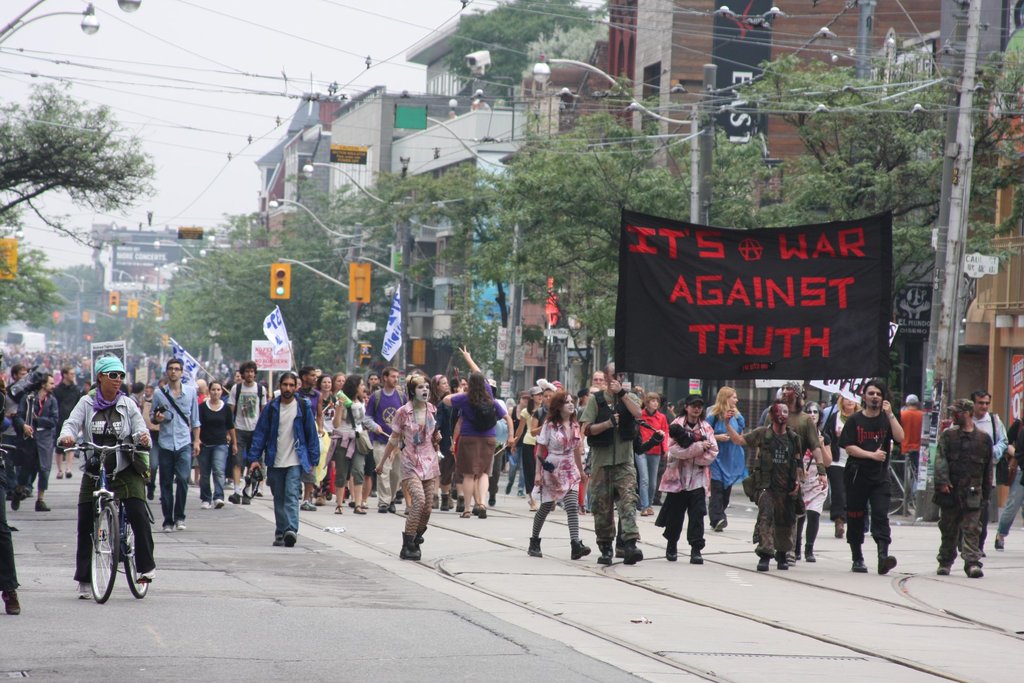 Eine Gruppe von Menschen, die eine Straße entlangmarschieren, Fahnen und Transparente schwingend, mit einer Person auf einem Fahrrad, umgeben von Laternenpfählen, Stromkabeln, Ampeln, Verkehrsschildern, Bäumen und Gebäuden.