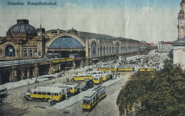 Schwarz-weiß-Postkarte vom Dresdner Hauptbahnhof: Eine belebte Straße mit Fahrzeugen, Passanten, Bäumen, Gebäuden und einem bewölkten Himmel.