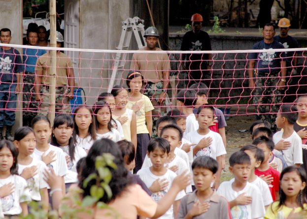 Eine Gruppe von Kindern vor einem Netz mit einer Frau im Vordergrund, während im Hintergrund Menschen mit Helmen, eine Leiter, eine Wand und Bäume zu sehen sind.