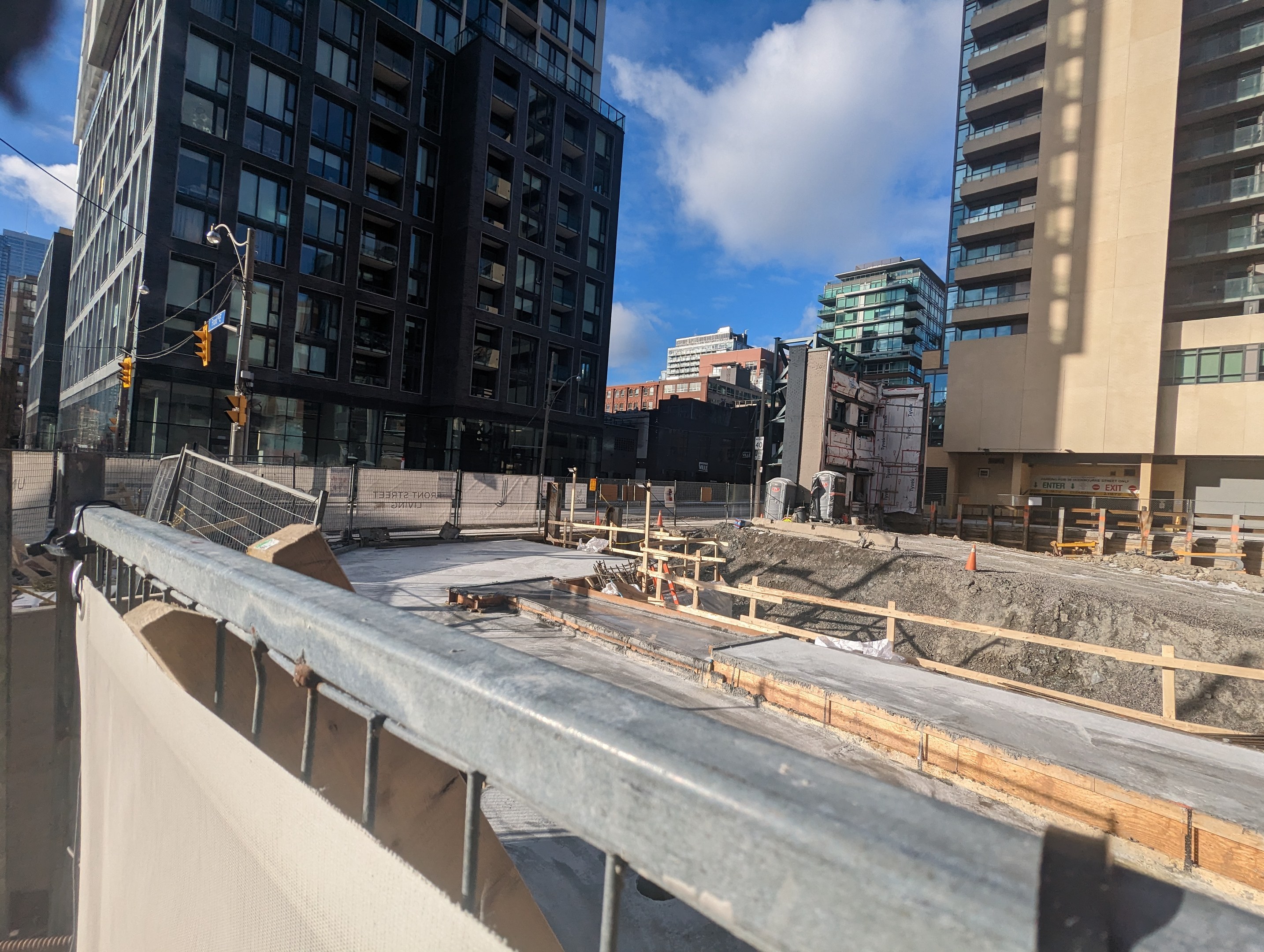 Baustelle in Toronto mit einem großen Gebäude im Bau, umgeben von Absperrungen, Pfählen, Verkehrsschildern und Beschilderung, unter einem bewölkten Himmel.