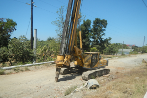 Bohrgerät an der Seite einer Schotterstraße mit Gras, Steinen, einem Rohr, Bäumen, Strommästen mit Drähten, einem Haus und einem bewölktem Himmel.