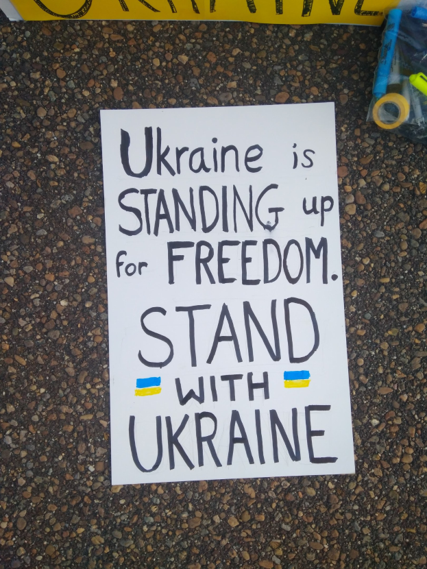 Ein weißes Blatt Papier mit dem Text "Ukraine steht für Freiheit auf, stellt Euch mit Ukraine" auf dem Boden vor einer gelben Fahne, mit einer Plastikhülle mit verschiedenen Gegenständen rechts.
