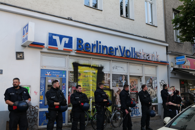 Eine Gruppe von Polizeibeamten steht vor einer Berliner Volksbank, einige halten Helme in den Händen, mit einem Auto auf der rechten Seite, einem Baum auf der linken Seite und Gebäuden mit Fenstern und Texttafeln im Hintergrund.