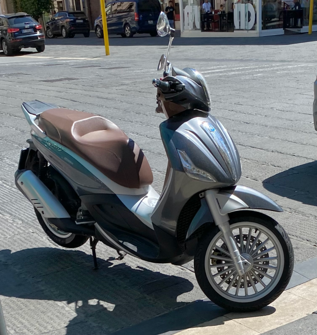 Ein Motorroller am Straßenrand geparkt mit einem Helm darauf, mit Gebäuden, Bäumen, Pfählen, Menschen, Stühlen, Tischen und anderen Gegenständen im Hintergrund.