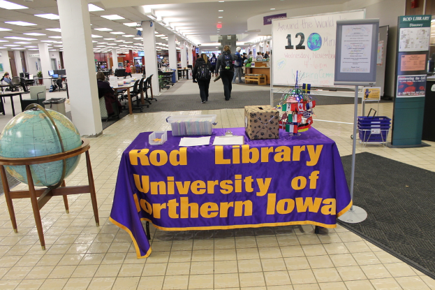 Eine Ausstellung mit einem Tisch, auf dem verschiedene Landesflaggen, eine Box, Papiere und ein Banner sowie links daneben eine Weltkugel, rechts daneben eine Anschlagtafel mit einigen Menschen in der Nähe und mehrere Tische und Stühle in der Ferne zu sehen sind.