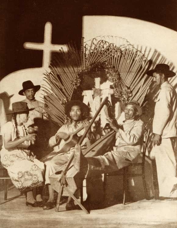 Schwarzes und weißes Foto einer Mariachi-Band, die vor einer Kirche sitzt, wobei einige Mitglieder Hüte tragen und Instrumente halten und ein Kreuz an der Wand im Hintergrund zu sehen ist.