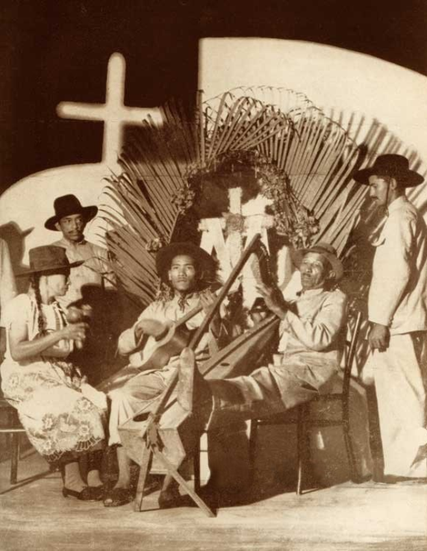 Schwarzes und weißes Foto einer Mariachi-Band, die vor einer Kirche sitzt, wobei einige Mitglieder Hüte tragen und Instrumente halten und ein Kreuz an der Wand im Hintergrund zu sehen ist.