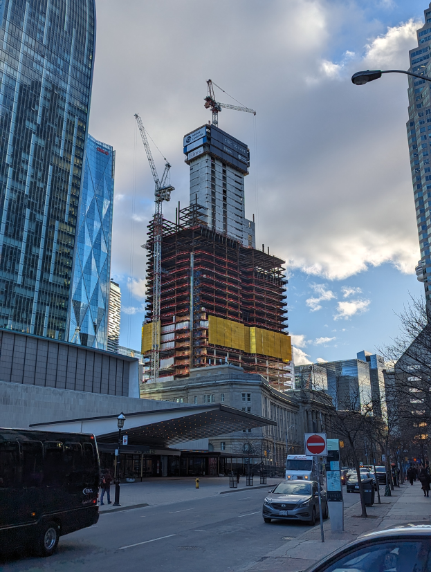 Eine belebte Stadtstraße mit Fahrzeugen, Fußgängern, Schildern, Laternen, Bäumen und im Hintergrund den One World Trade Center im Bau.