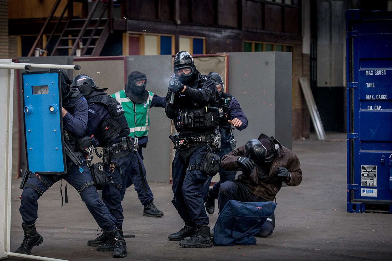 Polizeibeamte in Einsatzausrüstung stehen vor einem Gebäude in einer Reihe, einige halten Waffen, mit einer Person am Boden im Vordergrund und einem blauen Objekt mit Text auf der rechten Seite.
